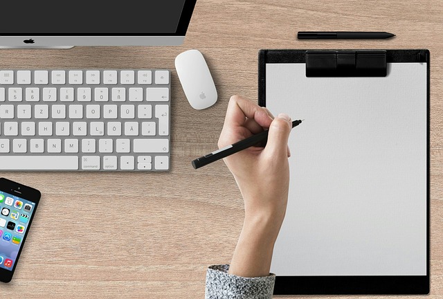 desk with computer, keyboard, monitor, mouse, phone, pen, and paper notebook